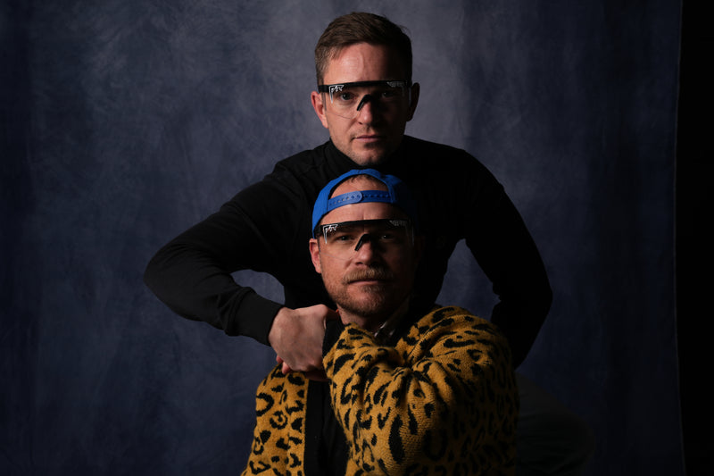 Two men wearing pit viper sunglasses and hats against a dark background
