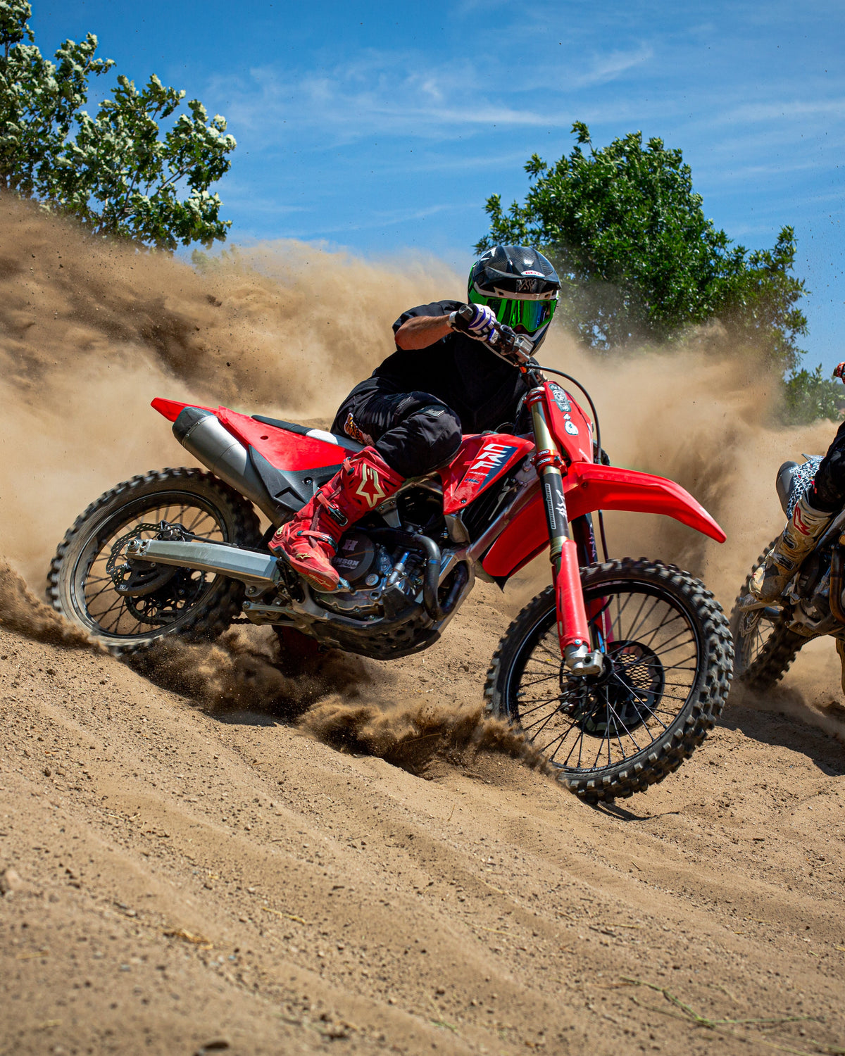 Two dirt bikers racing on a sandy track with trees in the background