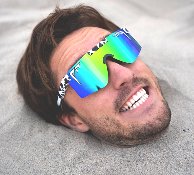 A man lying on sand wearing oversized shield-style sunglasses with mirrored blue-green lenses and white-and-black patterned arms.