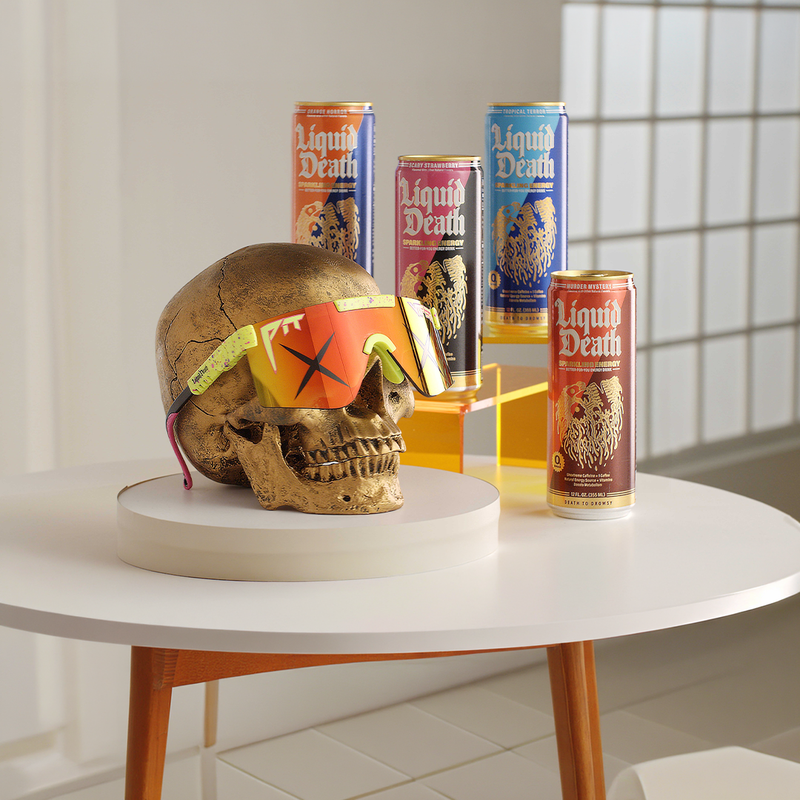 Skull with colorful pit viper sunglasses and 'Liquid Death' cans on a table