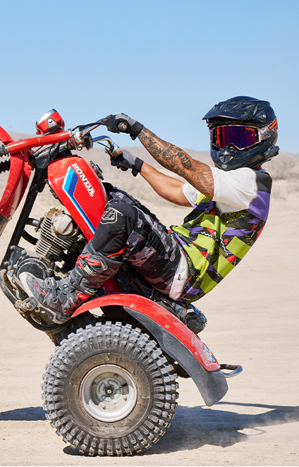 Person riding a red Honda dirt bike on a sandy surface with a clear blue sky.