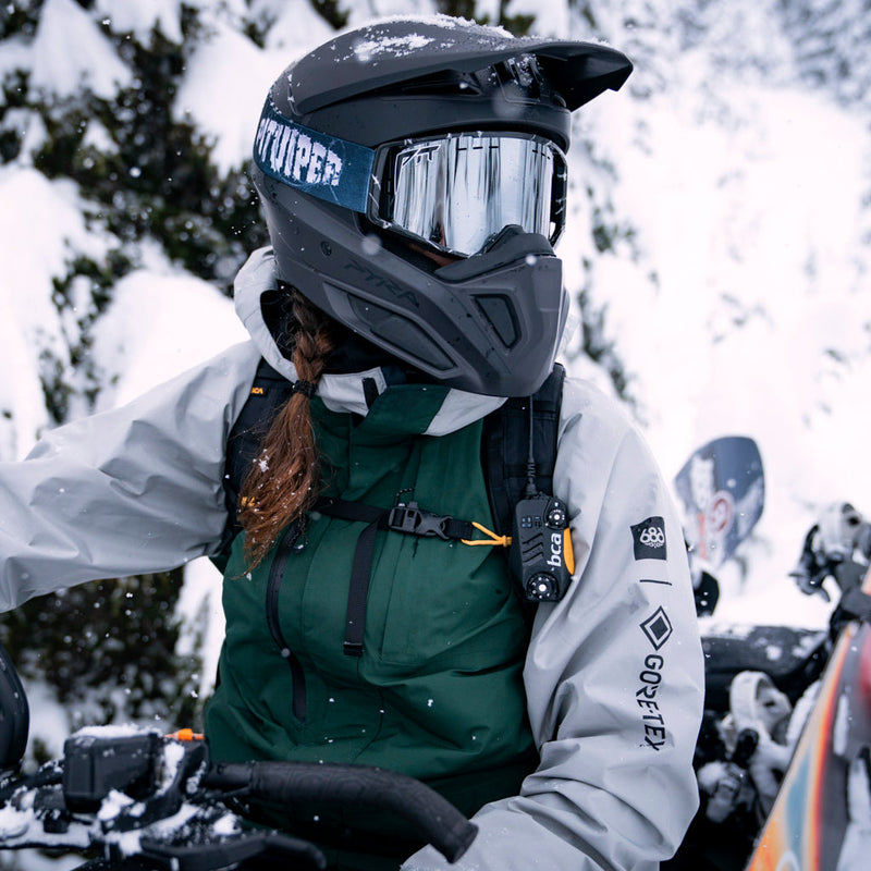 Silver | braaap.JPG | A person wearing a black full-face snowmobile helmet with a reflective visor, a dark green insulated jacket, and a light gray jacket, riding a snowmobile in a snowy outdoor setting.
