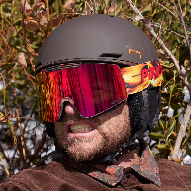  | pray_4_snow.JPG | A close-up of a person outdoors wearing a matte dark brown ski helmet and large mirrored ski goggles with a flame-patterned side strap. The background shows blurred branches and foliage.