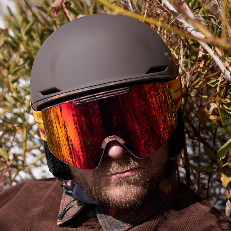 / Red | wheres_the_snow.JPG | A close-up of a person outdoors wearing a matte dark helmet and large ski goggles with a reflective red, orange, and yellow mirror finish. The background consists of blurred natural branches and foliage.