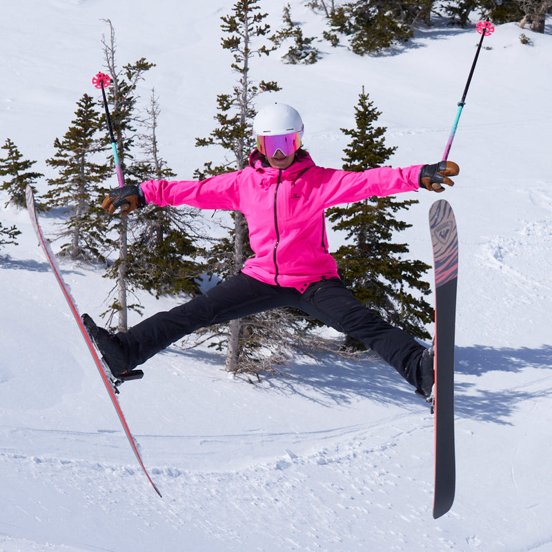 Pink | eagle.JPG | A skier jumps mid-air in a snowy mountain setting, wearing a bright pink ski jacket, black ski pants, a white helmet, and pink-tinted goggles, with ski poles and skis extended.