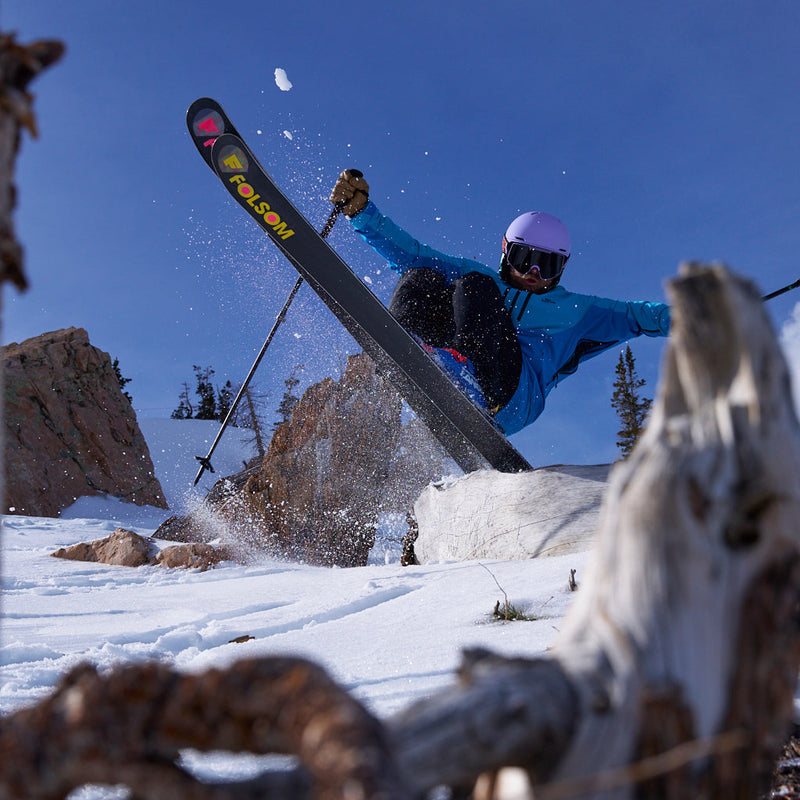 | tips_up.JPG | A skier jumps mid-air over snow with a rock wall and trees in the background. The skier wears a bright blue jacket, black pants, a light purple helmet, and goggles, and holds ski poles.