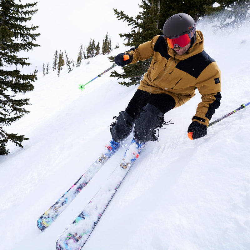  | how_much_wood_could_chuck_chuck.JPG | A skier carving through deep powder snow on a mountain, wearing a tan and black ski jacket, black pants, a dark helmet, and ski goggles, with evergreen trees in the background.