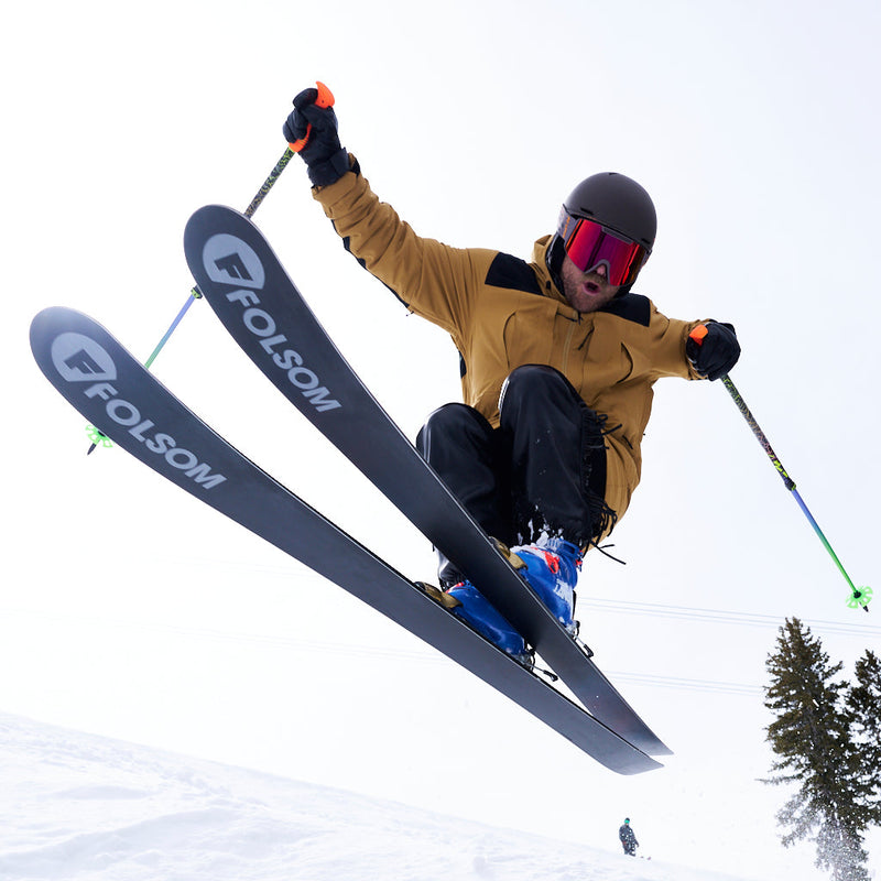 / Red | how_much_chuck_chuck.JPG | A skier mid-jump in a snowy outdoor setting, wearing a tan ski jacket, black pants, a dark helmet, and goggles, with Folsom skis and ski poles visible.