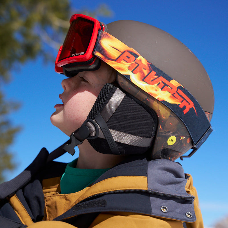  | smol_helmet.JPG | A close-up lifestyle shot of a child wearing a matte dark gray helmet with a flame-patterned strap, red ski goggles, and a yellow and navy winter jacket against a bright blue sky.