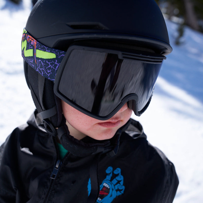 Smoke / Small | photo_of_person.JPG | A close-up of a child wearing a matte black ski helmet and black ski goggles with a purple and neon green patterned strap, set against a snowy outdoor background.