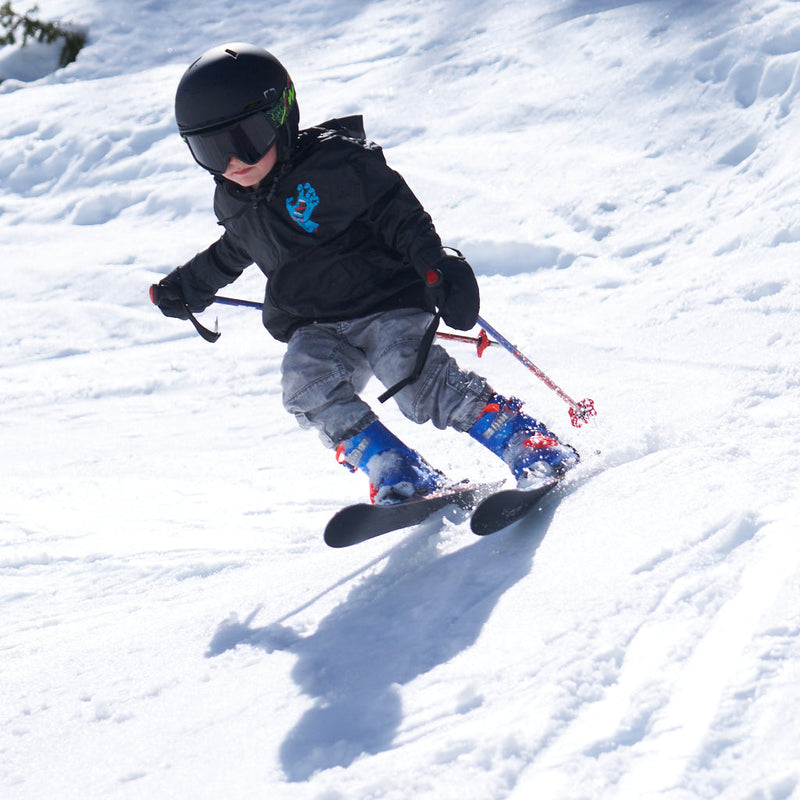Smoke / Small | photo_of_a_jean_skier.JPG | A child skier in a black helmet, black jacket, gray pants, and blue boots carving through fresh snow, holding ski poles.