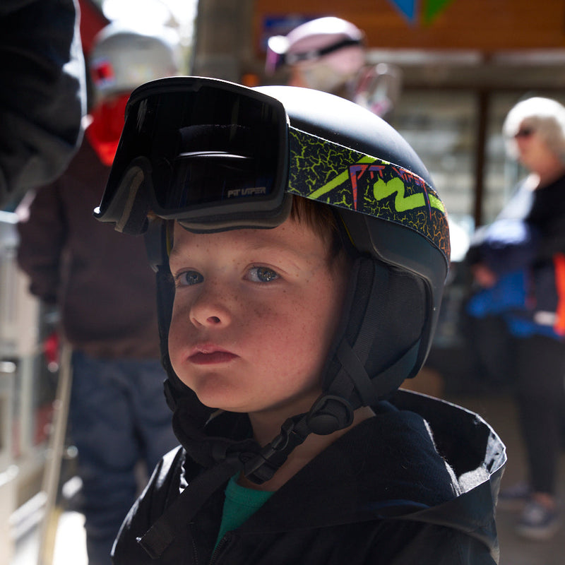 Smoke / Small | sup_dad.JPG | A close-up lifestyle shot of a young boy wearing a black ski helmet with goggles resting on his forehead and a colorful graphic band. He is dressed in a black ski jacket, set against a blurred outdoor background with other people and snow gear.