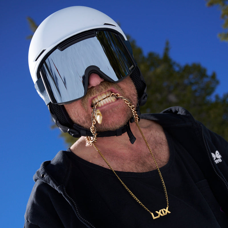 | photo_of_a_chuck.JPG | close-up lifestyle shot of a man wearing a white ski helmet and black goggles, wearing a gold chain necklace with a custom 'LXOX' pendant and a gold chain hanging from his teeth. The background shows a bright blue sky and green trees.
