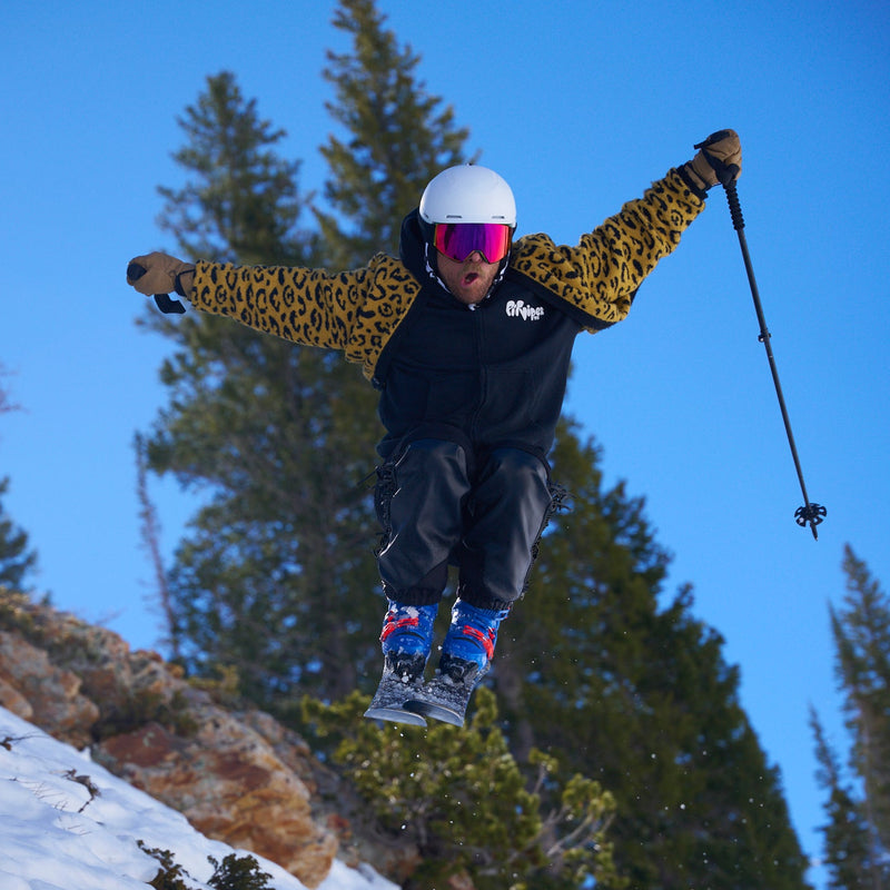 Photo of bearded man wearing a leopard print sweater, a white ski helmet and blue ski boots catching air while also wearing the BN 5000 Goggles from Pit Viper.
