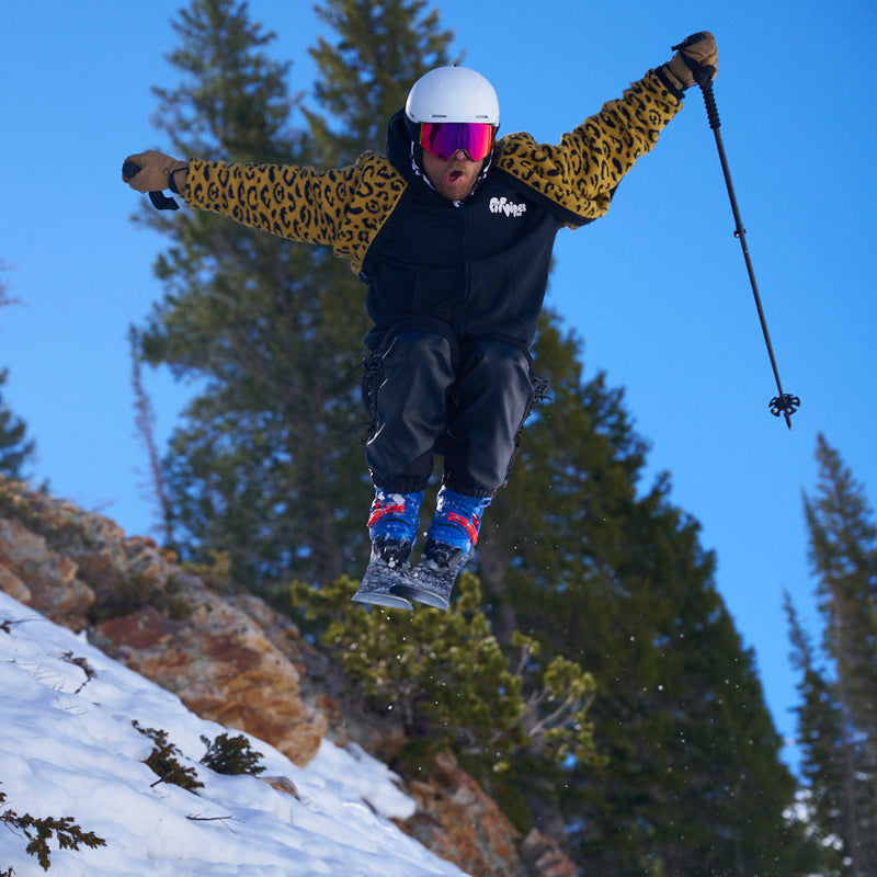 | big_air_chuck.JPG | A skier mid-jump in a snowy outdoor setting, wearing a black and leopard-print jacket, dark pants, a white helmet, and bright pink goggles, with trees and blue sky in the background.