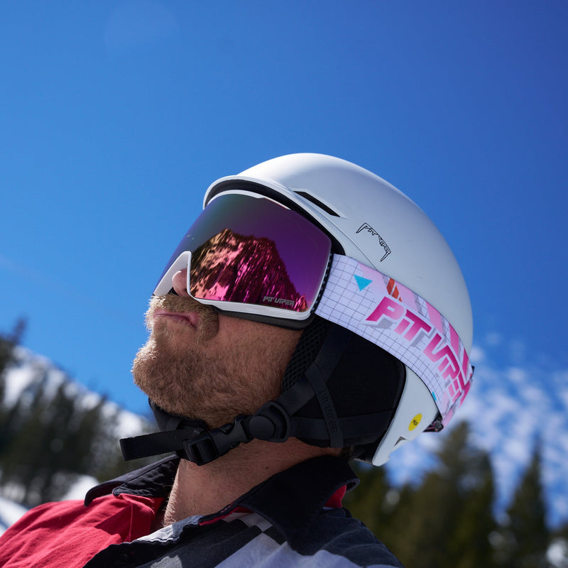 A close-up side profile of a person wearing a white ski helmet with pink mirrored goggles, set against a bright blue sky and snowy mountain background.