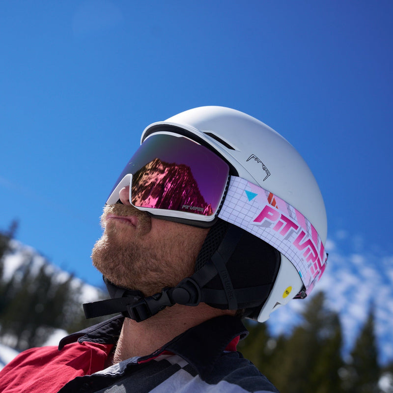 Pink | what.JPG | A close-up side profile of a person wearing a white ski helmet with pink mirrored goggles, set against a bright blue sky and snowy mountain background.