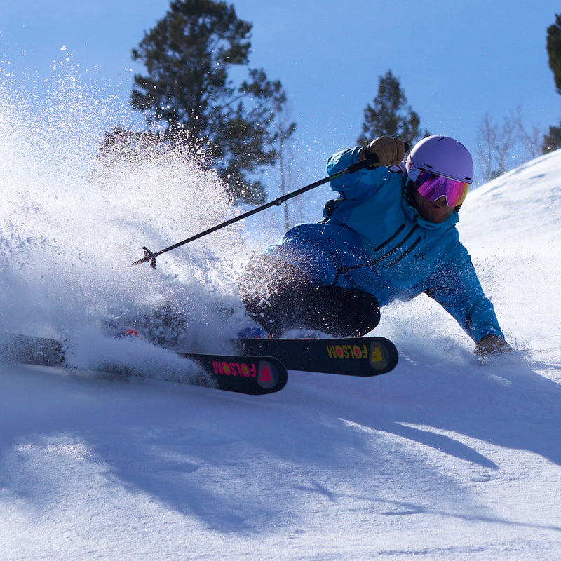| sprayer.JPG | A skier carving through fresh powder on a snowy slope, wearing a bright blue jacket, black pants, a light purple helmet, and pink-tinted goggles. The skier is captured mid-turn with snow spraying around them under a clear blue sky.