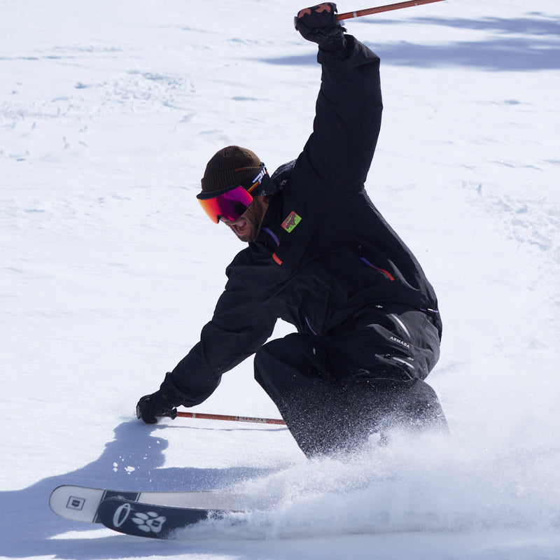 Red | slasher.JPG | A skier in a black ski jacket and pants, wearing orange-tinted goggles and a brown beanie, carving through fresh snow with ski poles and skis visible.