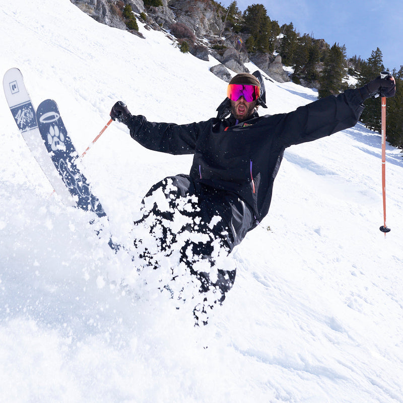 Red | ayyyy.JPG | A skier carving through deep powder snow on a steep mountain slope, wearing a black ski jacket, black pants, and bright pink-tinted goggles, with ski poles extended outward.