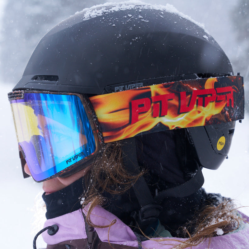 | photo_of_skier.JPG | A close-up side view of a person in a black ski helmet and goggles with a flame-patterned strap and blue mirrored lenses, wearing a light purple jacket in the snow.