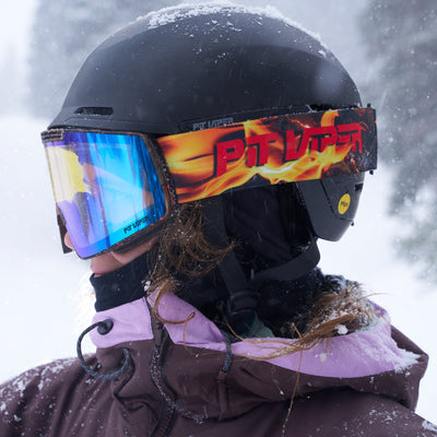 A person in a snowy outdoor setting wearing a black ski helmet, blue mirrored goggles with a red and yellow flame-pattern strap, and a brown jacket with a light purple collar.