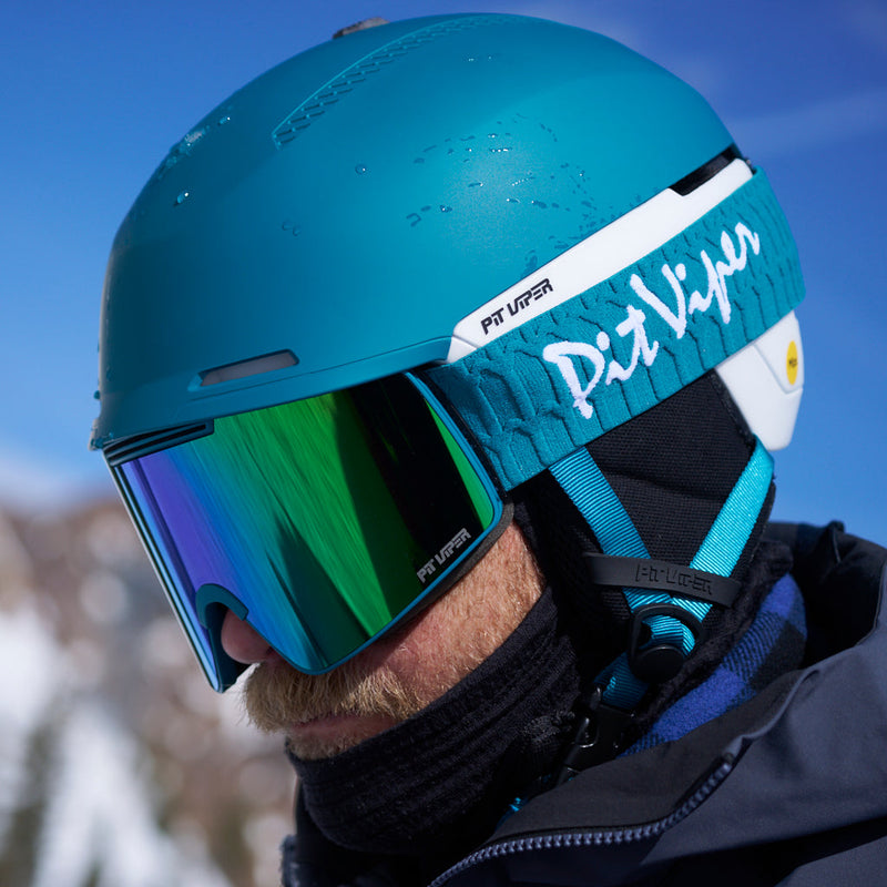 | photo_of_person.JPG | Close-up of a skier wearing a teal Pit Viper helmet with a matching teal and white head strap, reflective green-tinted goggles, and a black neck gaiter in a snowy outdoor setting.