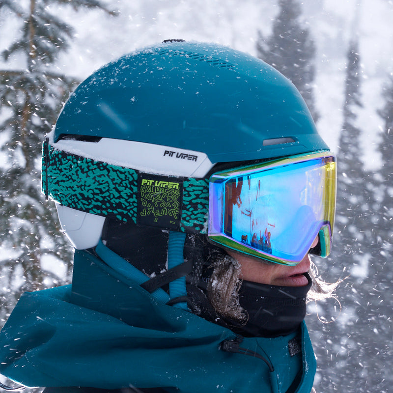 HDPV Smoke | photo_of_person.JPG | A close-up of a skier wearing a teal Pit Uper helmet, matching teal jacket, and reflective blue ski goggles with yellow edges, set against a snowy forest background with snow falling.
