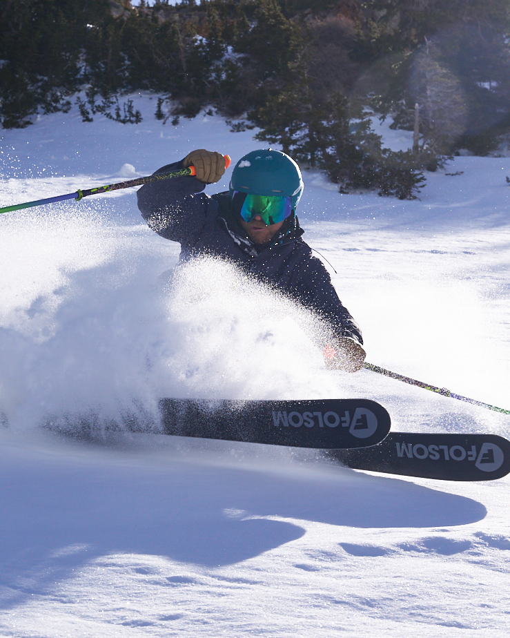 Man skiing making a strong turn in soft snow wearing a navy jacket with a teal helmet with matching pit viper goggles.