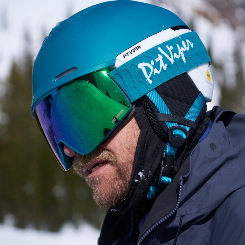 Blue-Green | photo_of_pro.JPG | A close-up of a person wearing a bright teal ski helmet with mirrored green and blue goggles, a white headband with teal branding, and a dark winter jacket in a snowy outdoor setting.