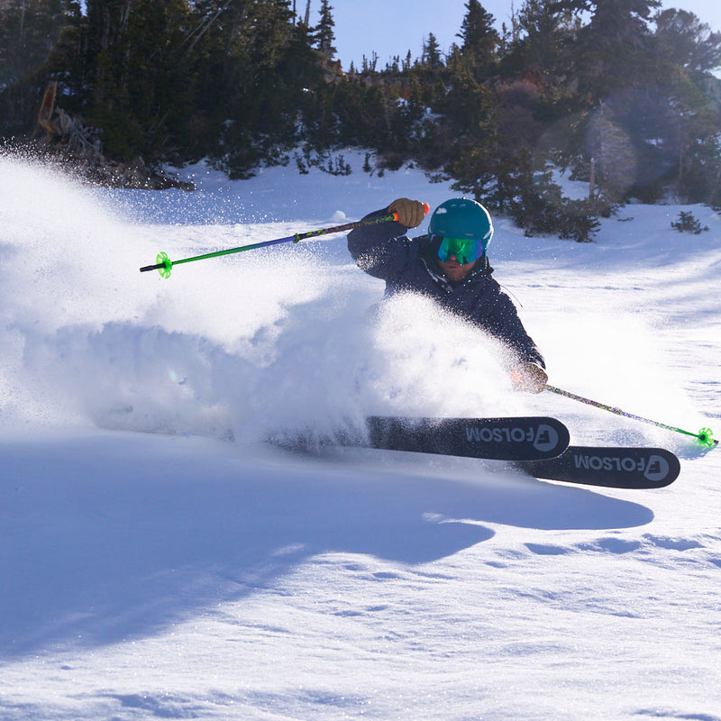 Blue-Green | spray.JPG | A skier carving through deep powder snow, creating a large spray of snow behind them. The skier is wearing a dark jacket, bright green goggles, and a teal helmet, with ski poles and skis visible.