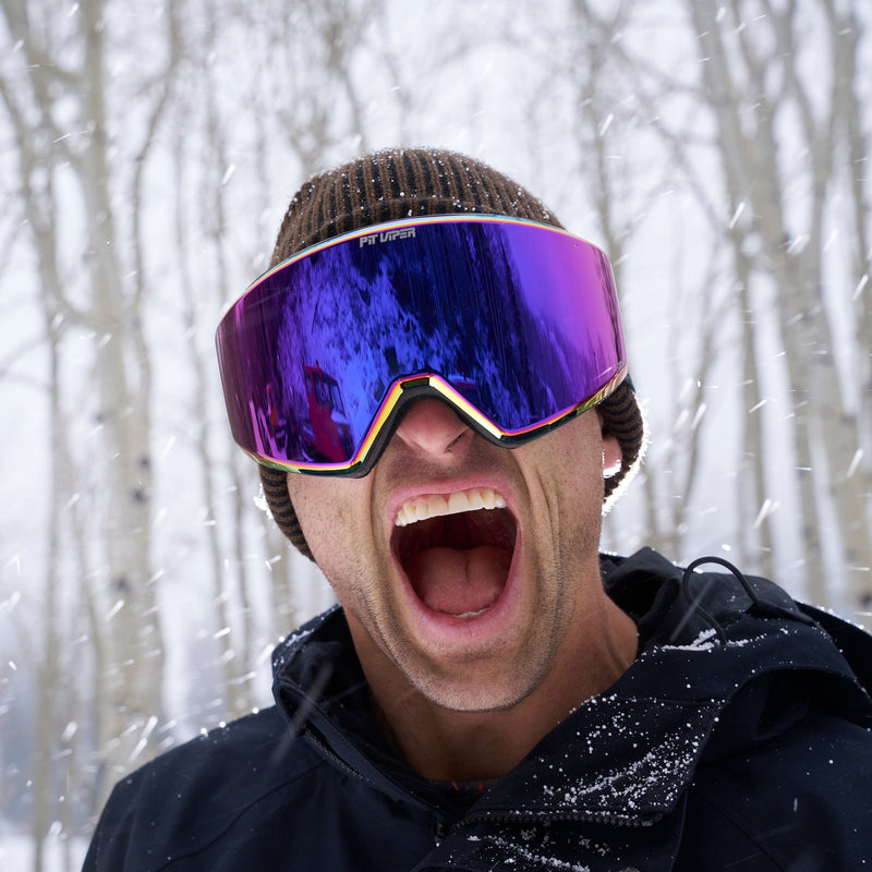 A man wearing a brown knit beanie and a black winter jacket shouts with his mouth wide open while wearing large ski goggles with a purple and blue mirrored lens, set against a snowy background with bare trees.