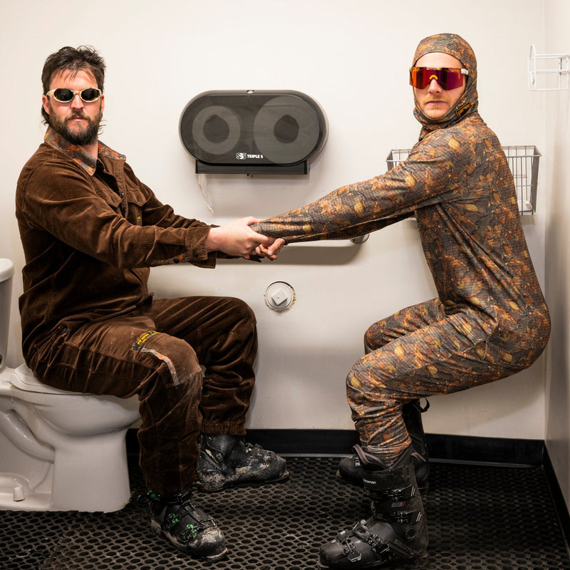 | so_cute_and_fun.JPG | Two men in full-body camouflage suits pose inside a small white bathroom, holding hands. One man sits on a toilet wearing a brown velour two-piece outfit and sunglasses, while the other stands crouching in a brown and tan camouflage full-body suit with red-tinted sunglasses.