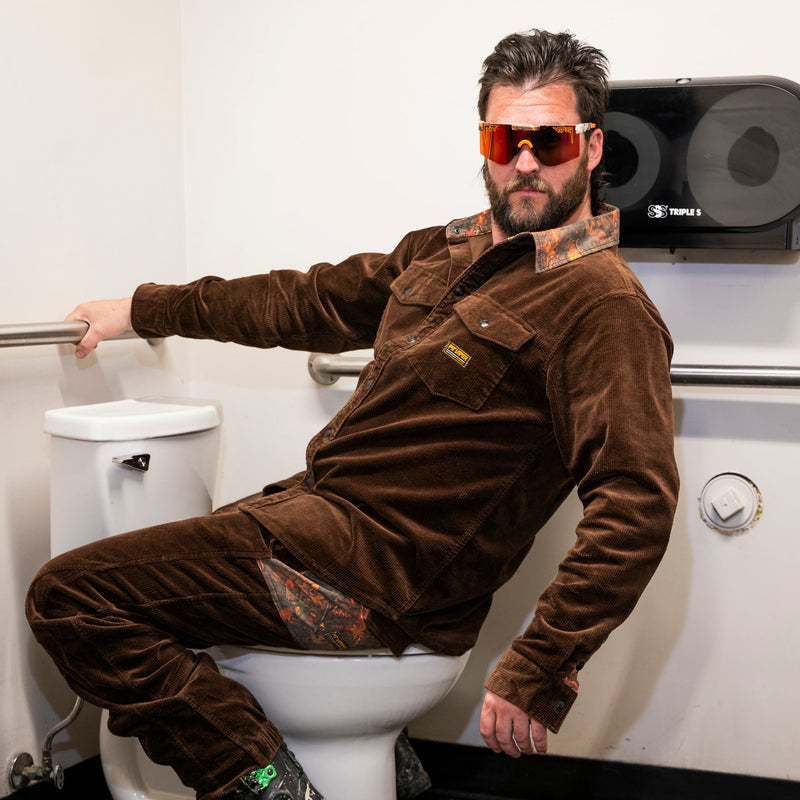 | toilet_man.JPG | A man wearing a brown corduroy jumpsuit and patterned sunglasses sits on a white toilet in a bathroom setting.