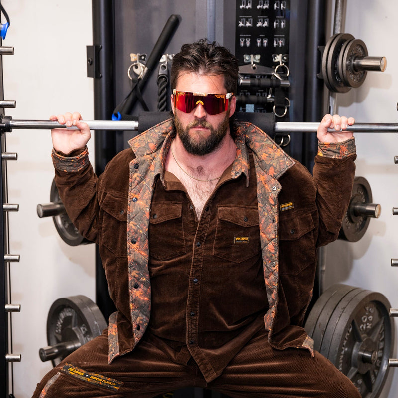| photo_of_strong_person.JPG | A man in a brown corduroy tracksuit and orange-tinted sunglasses sits inside a power rack in a gym setting.