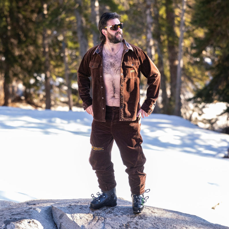 | photo_of_person.JPG | A man wearing a brown corduroy overshirt, brown pants, and black boots stands on a large rock in a snowy forest setting.