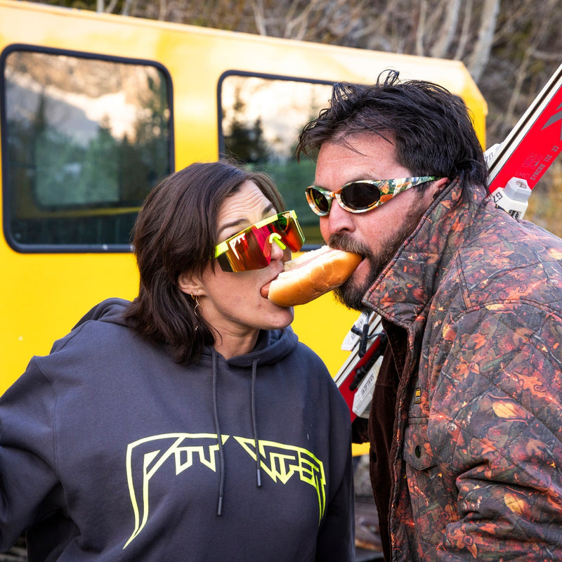 | photo_of_person.JPG | A man and a woman outdoors in front of a yellow vehicle, with the woman wearing a dark hoodie with a neon yellow logo and the man wearing a camouflage-patterned jacket. Both are holding skis, and the man is eating a hot dog.