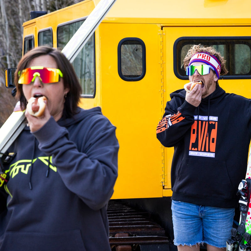 | photo_of_people.JPG | Two people wearing ski goggles and hoodies eat hot dogs in front of a bright yellow snowmobile in an outdoor winter setting.