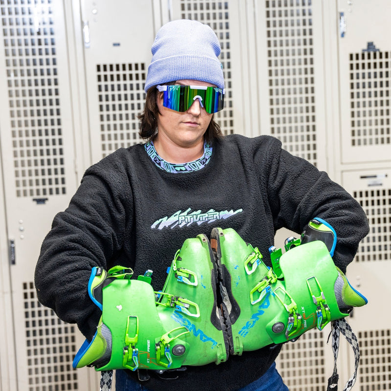 | photo_of_person2.JPG | A person wearing a light blue beanie, mirrored ski goggles, a black sweatshirt, and blue jeans holds a pair of bright neon green ski boots in front of a row of beige metal lockers.