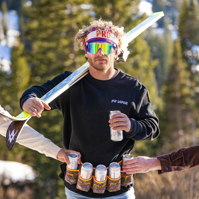 | man_holding_only_skis.JPG | A man outdoors in a snowy mountain setting, wearing a black sweatshirt with 'FIT VAPOR' on the chest, blue denim shorts, and holding a ski over his shoulder. He is surrounded by hands holding energy drinks and a can, with pine trees in the background.