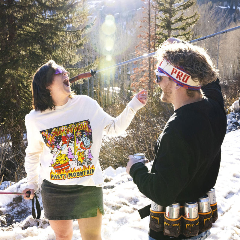 | hot_dog.JPG | Two people outdoors in a snowy forest setting, each holding ski poles. The person on the left wears a white graphic sweatshirt and dark skirt, while the person on the right wears a black jacket and a white headband with red text.