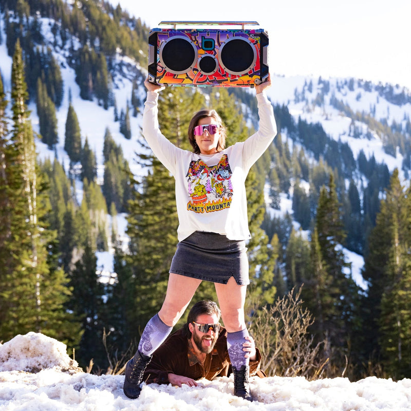 | play_freebird.JPG | A woman in a snowy mountain setting holds up a large, brightly colored boombox-style speaker above her head while lying a man on the snow below her. She is wearing a white graphic long-sleeve shirt, a dark gray mini skirt, and knee-high socks.