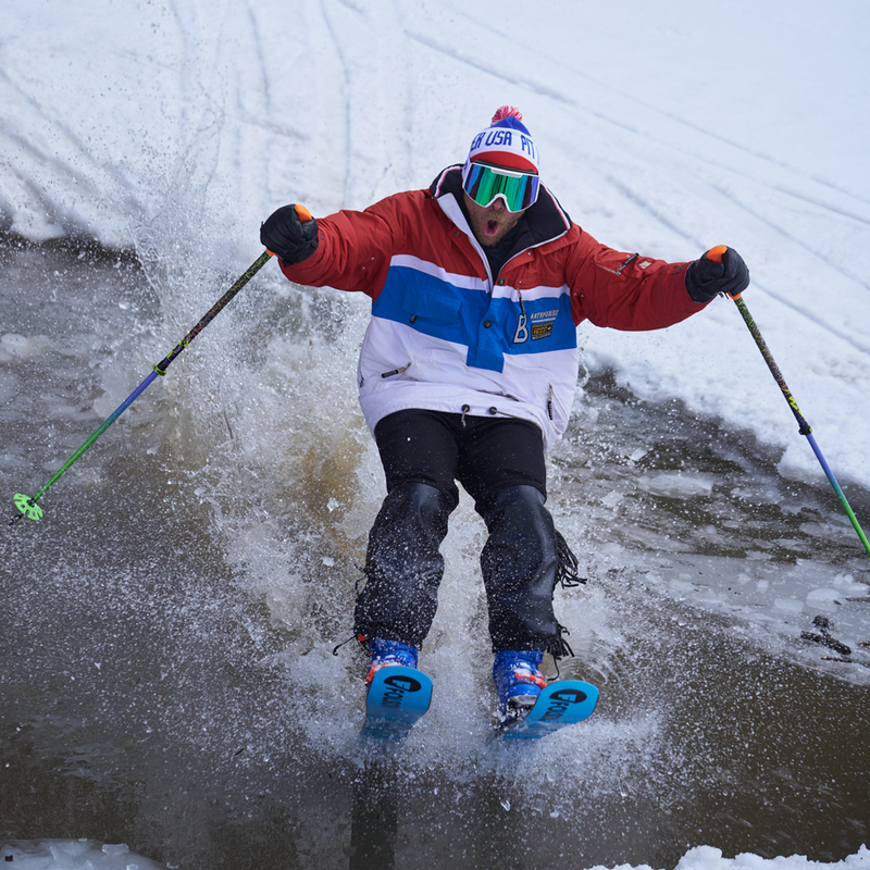 Blue-Green / Small | skimmah.JPG | A skier in a red, white, and blue ski jacket, black pants, and blue skis glides through splashing water on a snowy slope, holding ski poles.