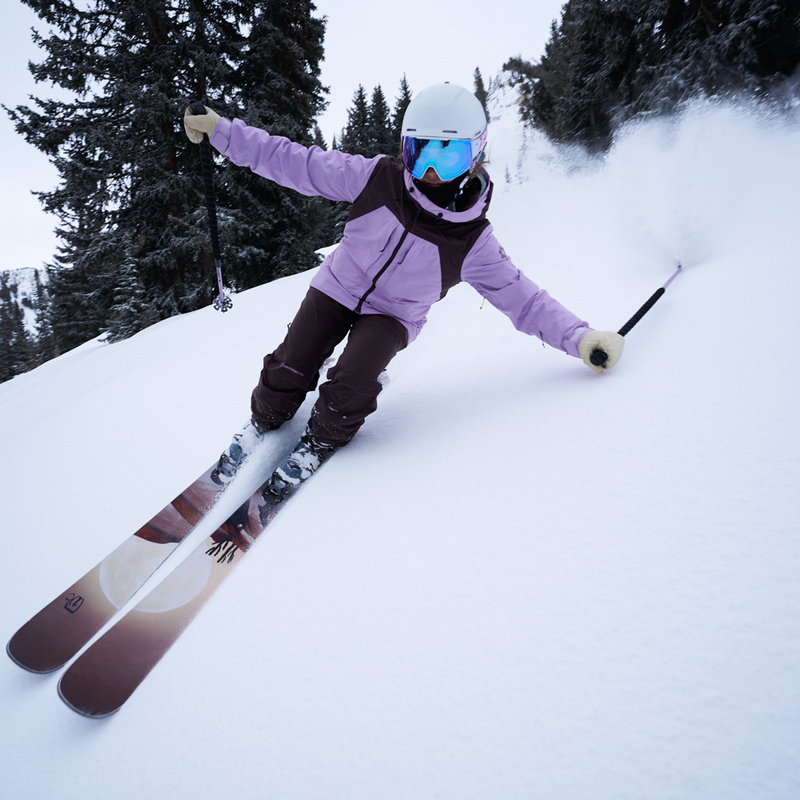 | photo_of_skier.JPG | A skier carving through deep snow on a steep slope, wearing a light purple jacket, dark brown pants, a white helmet, and blue mirrored goggles. Snow spray is kicked up behind them, with evergreen trees in the background.