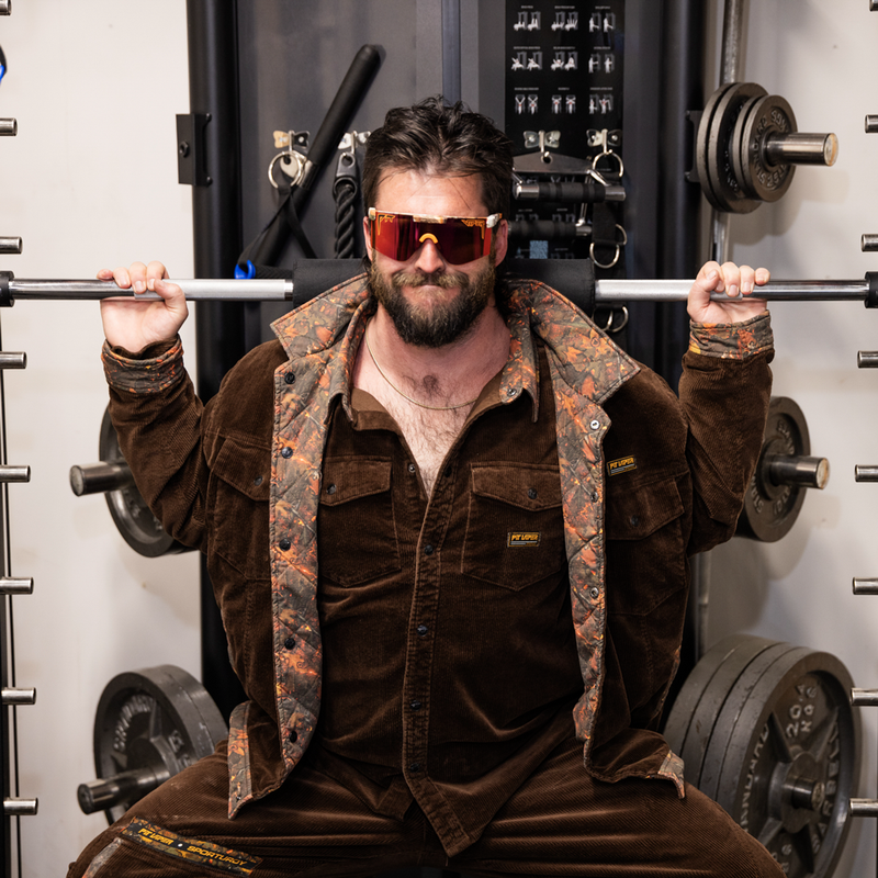 | buff.JPG | A man in a gym wearing a brown corduroy button-up shirt and matching brown corduroy pants, featuring a patterned lining on the collar and cuffs. He is wearing reflective orange-tinted sunglasses and posing behind a barbell on a weight rack.